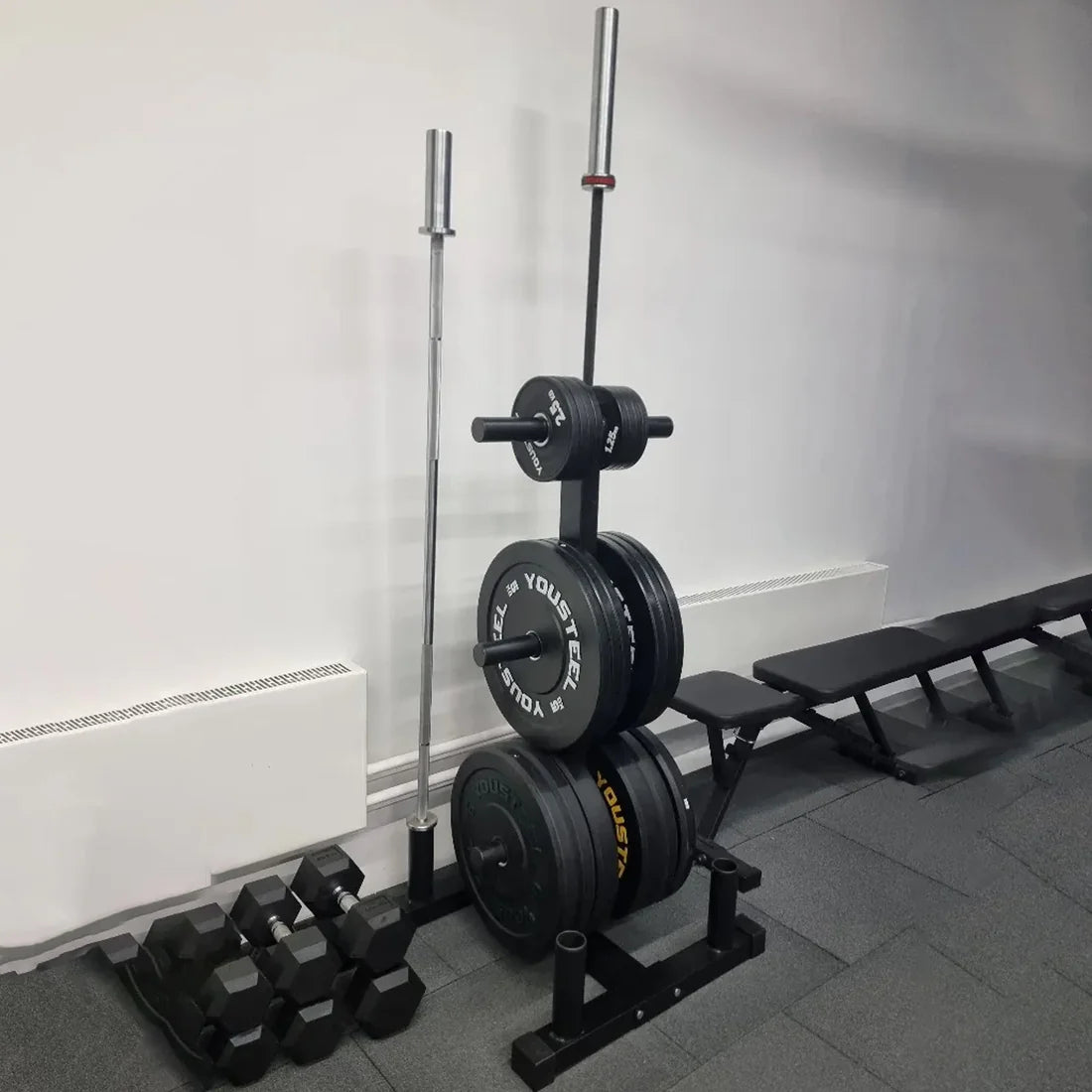 Gym equipment including weights and dumbbells against a white wall.