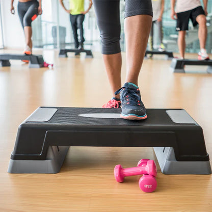 Person using an exercise step with pink dumbbells on a wooden floor in a gym setting